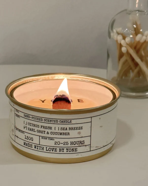 Lit Daily Reminder Candle with wooden wick flame burning in gold tin beside glass jar with matches