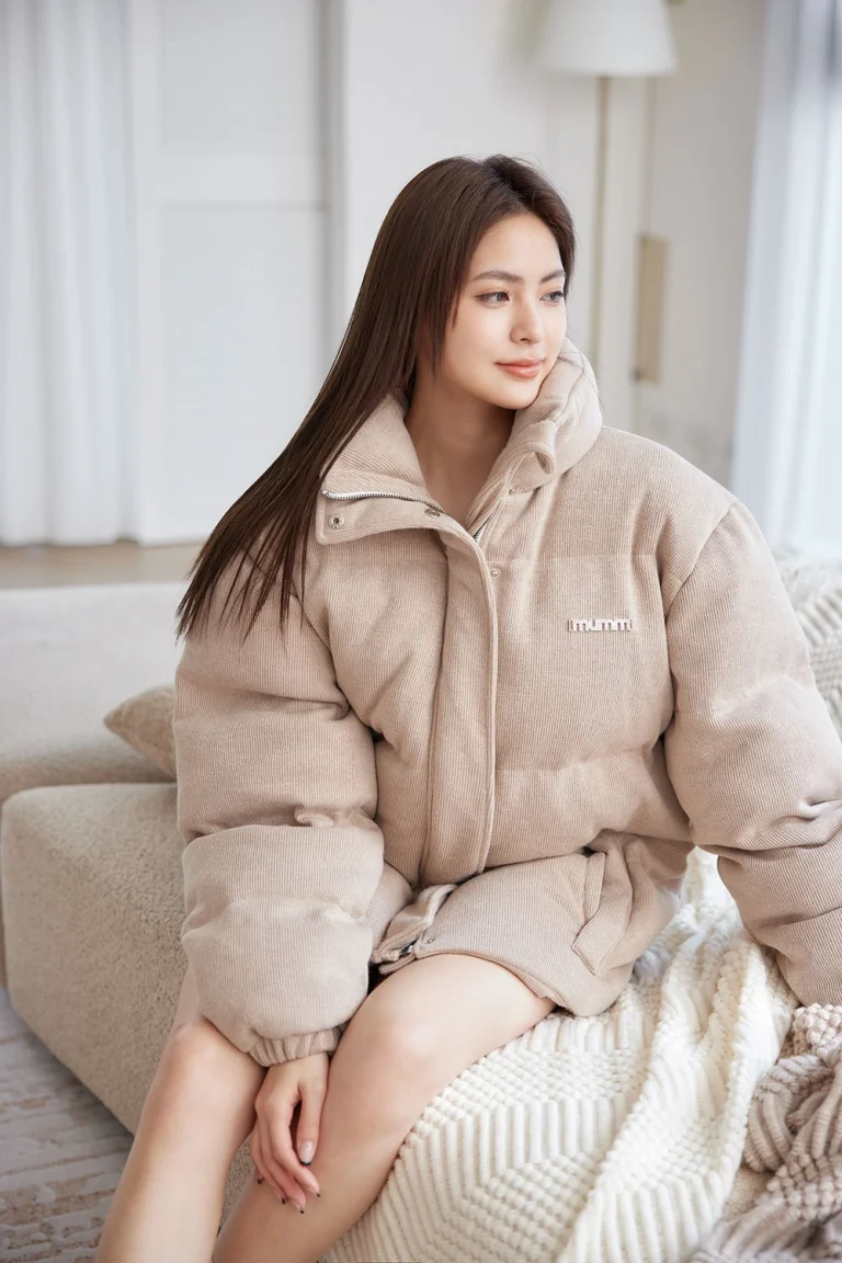 Woman wearing beige thermal puffer jacket relaxing on neutral sofa with white curtains in cozy living room